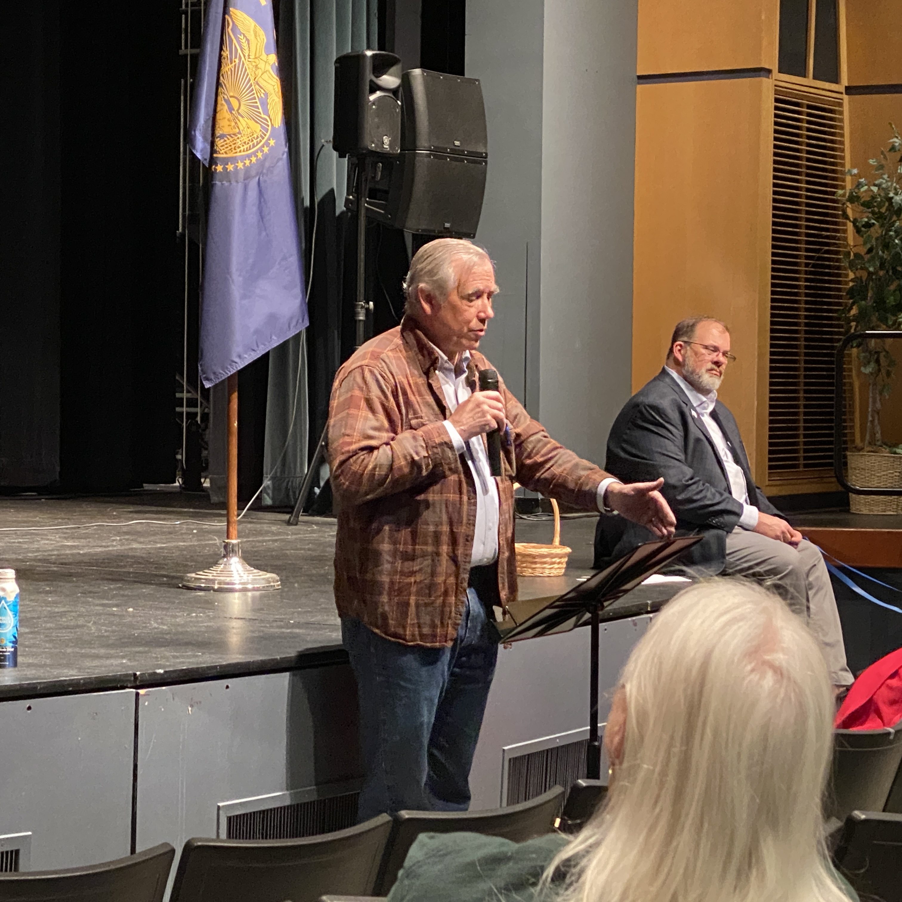 A theater auditorium. A man, Jeff Merkley, speaks to the audience with a microphone